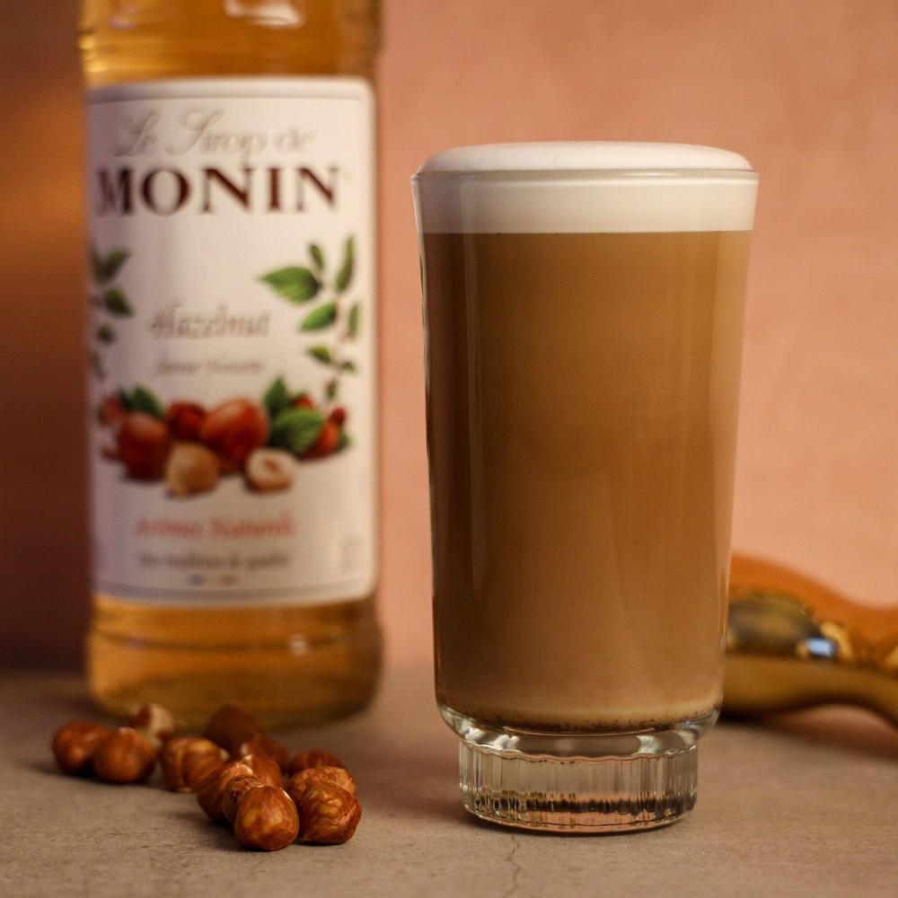 Monin Hasselnøtt sirup 70 cl flaske ved siden av en latte i glass med hasselnøtter.
