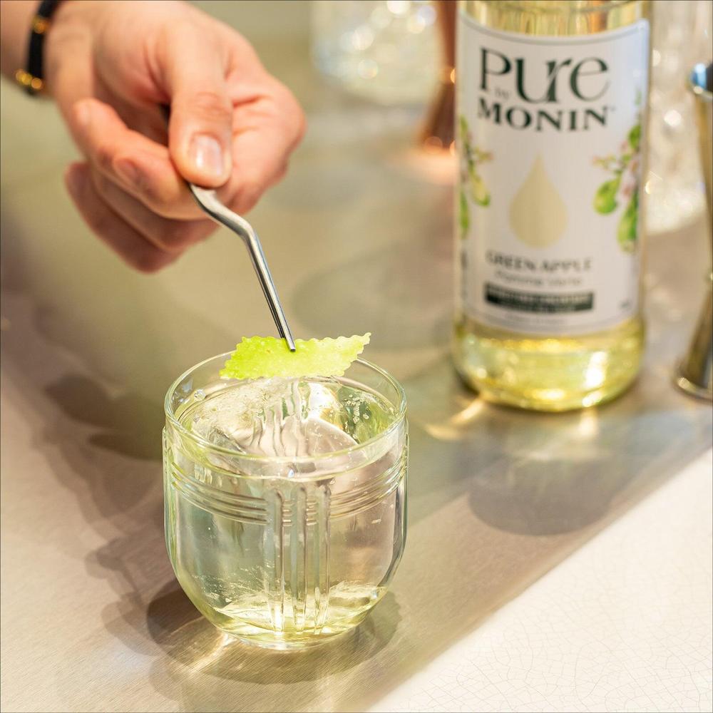 Pure by Monin Green Apple sirupflaske ved siden av en hånd som pynter en drink med lime.