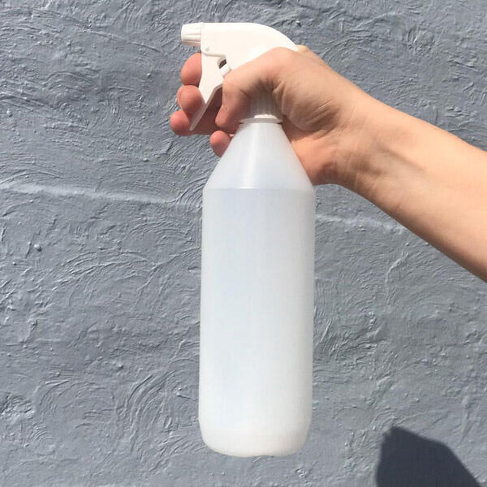 Hvit 1 liters spruteflaske med trigger, holdt i en hånd mot en grå vegg.
