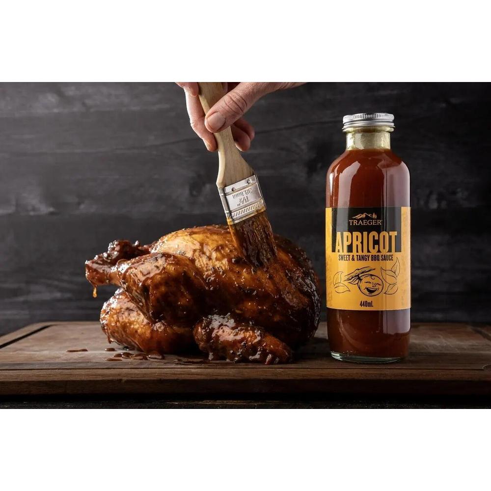 Traeger Apricot BBQ-saus 440ml flaske ved siden av penslet grillet kylling på et skjærebrett.