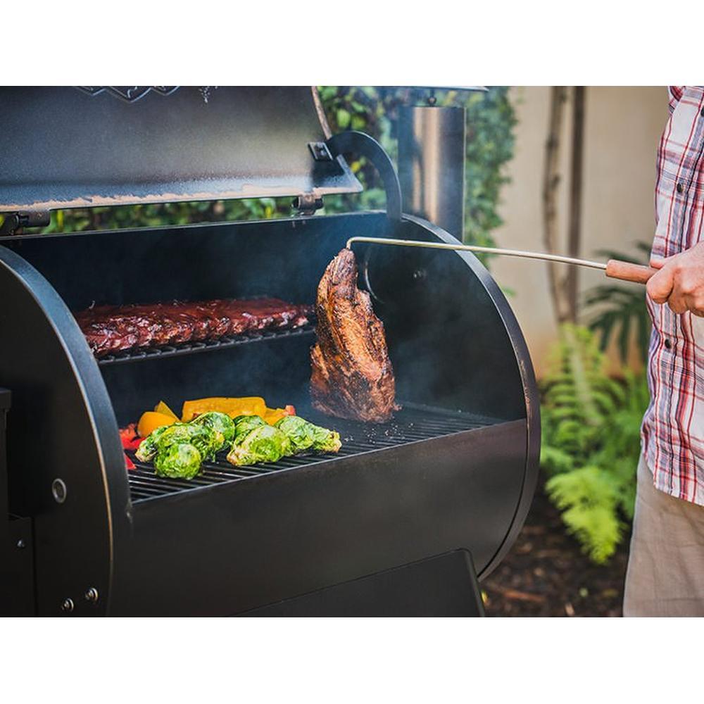 Traeger BBQ Pig Tail vendekrok i rustfritt stål med teak-håndtak snur kjøtt på en svart grill.