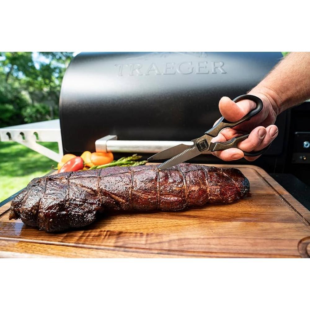 Traeger BBQ kjøttsaks med titanbelagte blader kutter grillet kjøtt på et skjærebrett, med grill i bakgrunnen.