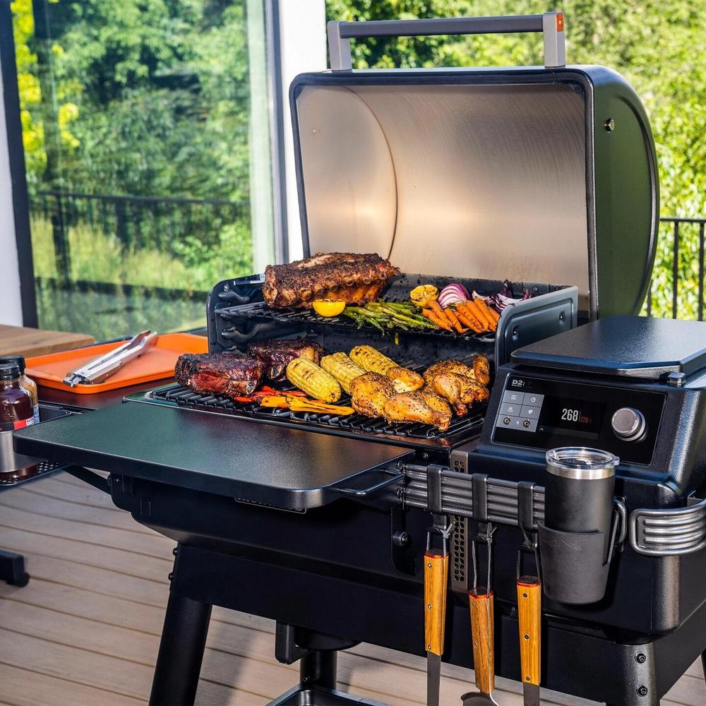 Traeger Ironwood pelletsgrill i svart med åpent lokk, grillet kjøtt og grønnsaker, verktøy og drikkekopp.