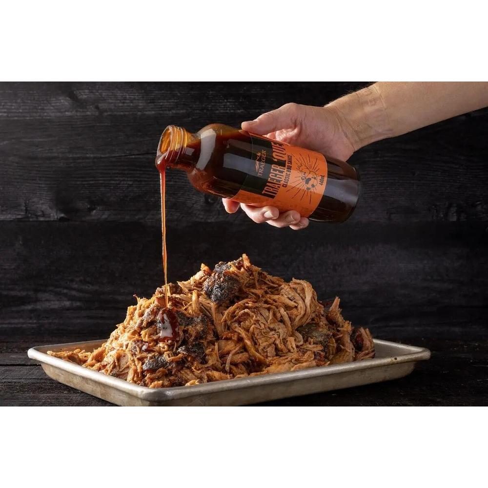 Traeger 'Que BBQ-saus helles over en stor porsjon pulled pork på et metallfat.
