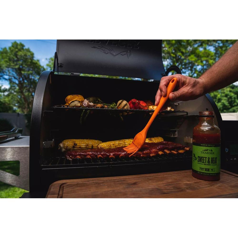 Oransje Traeger silikonpensel påfører marinade på grillribbe, med grill og sausflaske i bakgrunnen.